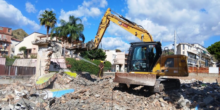 Rental Caterpillar CAT 313 GC crawler excavator by Alayan Rentals with hydraulic crusher engaged in a demolition site with dust suppression.