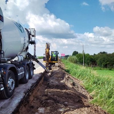 Rental crawler excavator and concrete mixer truck by Alayan Rentals engaged in concrete pouring for roadside trenching.