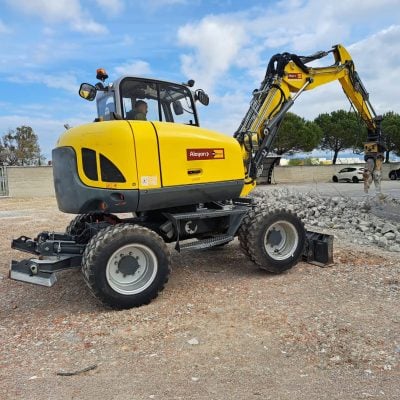 Rental compact wheeled excavator by Alayan Rentals driven by an operator on an excavation and land preparation site during the removal of a reinforced concrete wall.