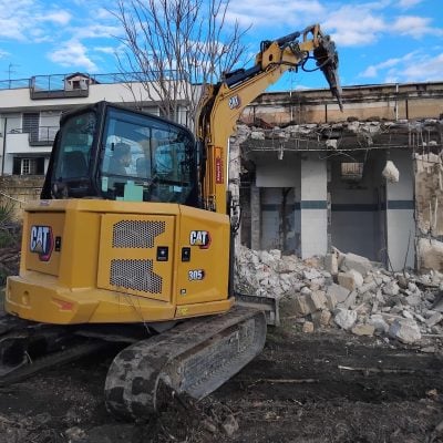 Rental Caterpillar CAT 305 mini crawler excavator by Alayan Rentals equipped with a hydraulic breaker on a building demolition site.