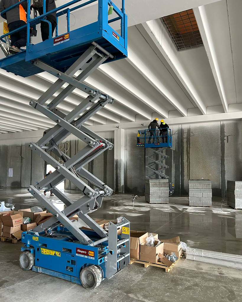 Due piattaforme aeree verticali a forbice elettriche blu a noleggio Alayan Rentals al lavoro all'interno di un capannone Alayan Noleggio