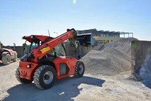 Red Manitou compact telehandler with raised aggregate bucket in a construction yard for rent - Alayan Rentals