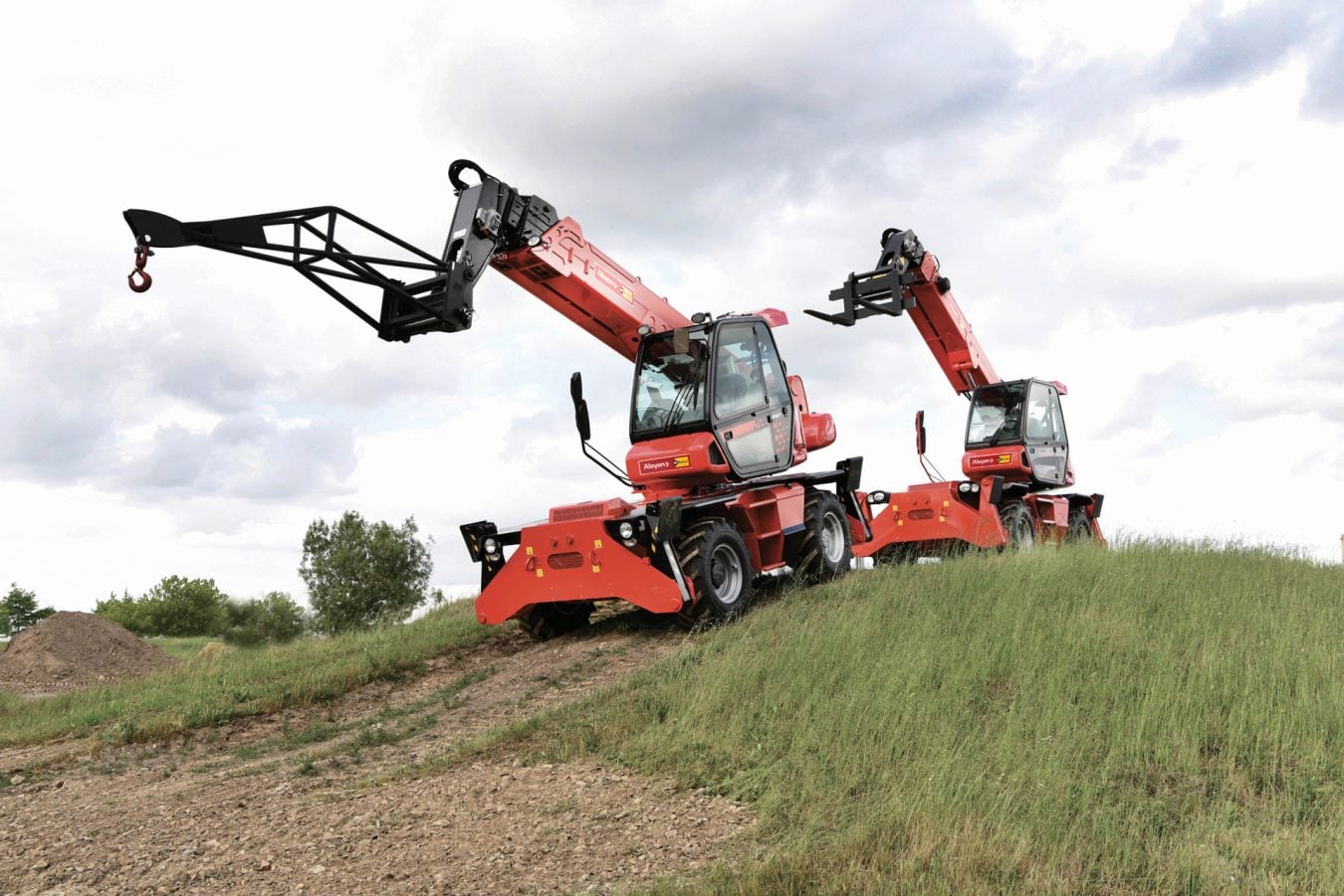 Due sollevatori telescopici rotativi Manitou rossi a noleggio Alayan Rentals, con falcone tralicciato e forche, su un pendio erboso Alayan Noleggio