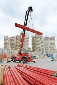 Roter drehbarer Manitou Teleskopstapler stabilisiert beim Heben eines Rohrbündels mit Winde auf Baustelle zur Miete - Alayan Rentals